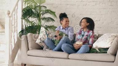 两个混血卷发女孩朋友坐在沙发上看<strong>喜剧电影</strong>，吃爆米花。 有趣的姐妹